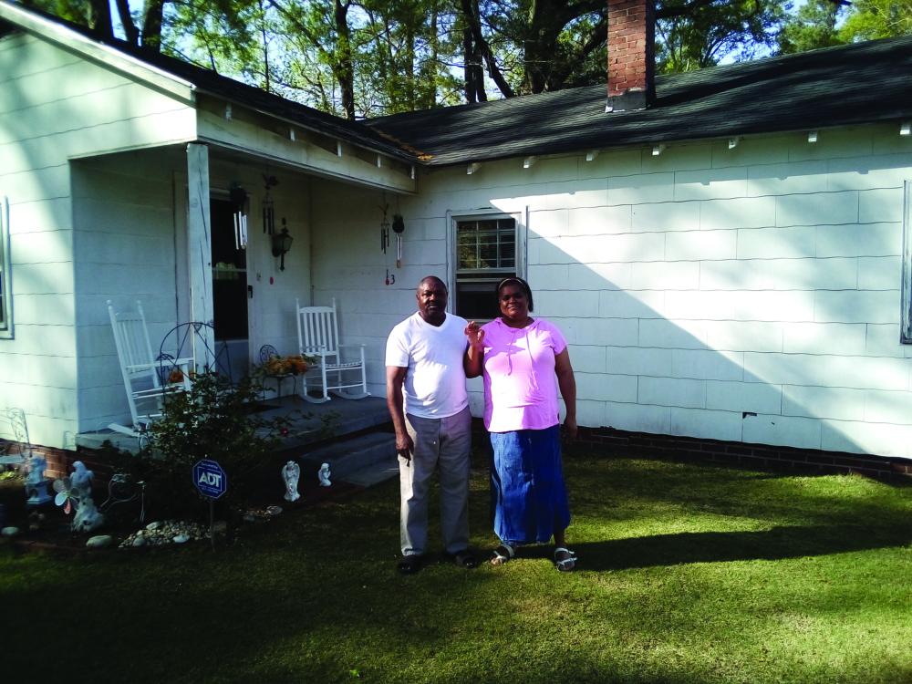 Two people stand in front of a white house with a porch.